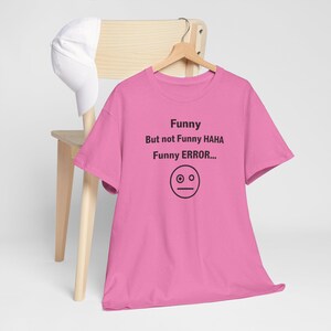 Puede incluir: Una camiseta rosa con el texto "Funny But not Funny HAHA Funny ERROR..." y un gr&aacute;fico de cara simple. La camiseta est&aacute; colgada en una percha de madera, con una gorra de b&eacute;isbol blanca y una silla de madera en el fondo.