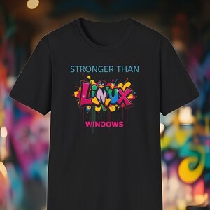 Könnte beinhalten: Schwarzes T-Shirt mit dem Text "STRONGER THAN" über einem farbenfrohen Graffiti-Design mit "LINUX" und einem Pinguin, sowie "WINDOWS" darunter. Der Hintergrund ist eine verschwommene Graffiti-Wand.