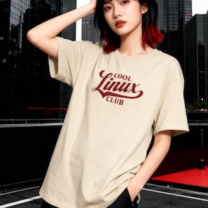 Peut inclure: T-shirt beige avec l'inscription "COOL Linux CLUB" en script bordeaux. Le t-shirt a des manches courtes et un col rond. L'image est prise en extérieur avec une vue sur la ville.