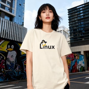 Può includere: T-shirt color crema con un pinguino dei cartoni animati e la scritta "Linux" in nero e giallo. La persona indossa la maglietta davanti a uno sfondo urbano con graffiti e edifici moderni.