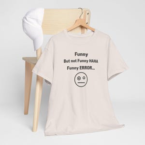 Puede incluir: Una camiseta beige claro con el texto "Funny But not Funny HAHA Funny ERROR..." y una cara neutral. Una gorra de b&eacute;isbol blanca descansa sobre una silla de madera detr&aacute;s de la camiseta.