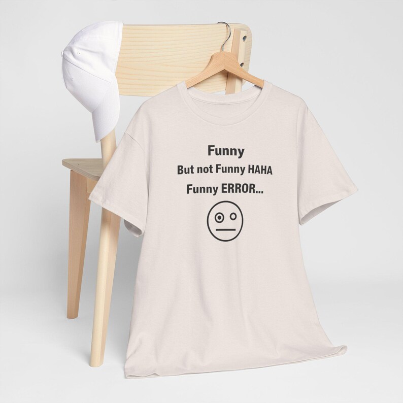 Puede incluir: Una camiseta beige claro con el texto "Funny But not Funny HAHA Funny ERROR..." y una cara neutral. Una gorra de b&eacute;isbol blanca descansa sobre una silla de madera detr&aacute;s de la camiseta.