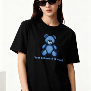 May include: Black t-shirt with a blue teddy bear design and the text "Your password is weak." The teddy bear has a light blue body and dark blue details. The shirt has short sleeves and a crew neck.