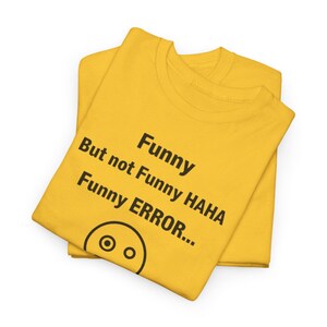 Puede incluir: Dos camisetas amarillas apiladas. La camiseta superior tiene el texto negro "Funny But not Funny HAHA Funny ERROR..." y un sencillo gr&aacute;fico de carita sonriente.