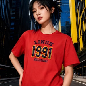Può includere: Una maglietta rossa girocollo con la scritta "LINUX 1991 HELSINKI" in blu navy e oro. La maglietta a maniche corte è realizzata in materiale morbido. L'immagine è scattata all'aperto.