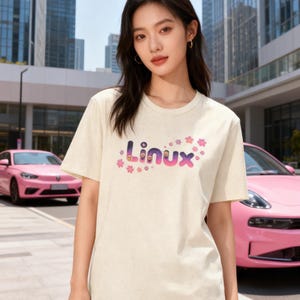 Peut inclure: Un t-shirt crème avec le mot "Linux" en dégradé de violet et rose, orné de motifs de fleurs roses. Le t-shirt à manches courtes est porté par une personne à l'extérieur, avec des voitures roses et des bâtiments en arrière-plan.