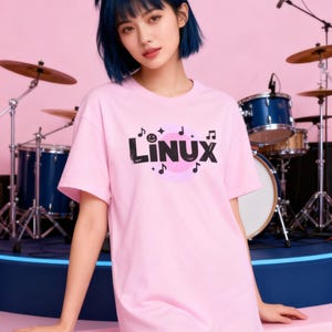 Könnte beinhalten: Ein hellrosa T-Shirt mit dem Wort "Linux" in Schwarz, mit Noten und einer rosa Grafik. Das T-Shirt wird von einer Person mit blauen Haaren vor einem Schlagzeug getragen. Der Hintergrund ist rosa.