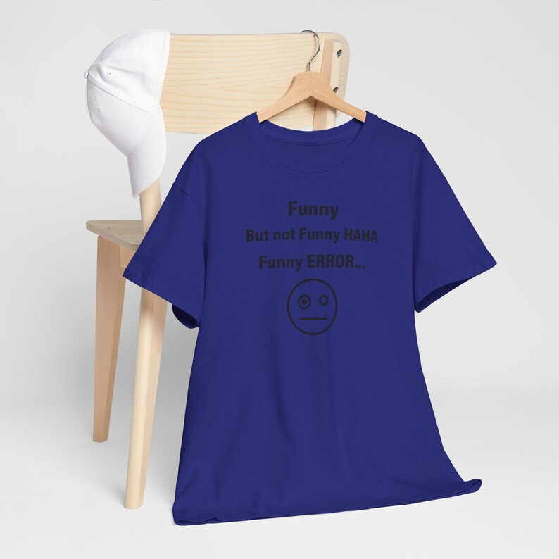 Puede incluir: Una camiseta azul real con el texto "Funny But not Funny HAHA Funny ERROR..." y un gr&aacute;fico de cara simple. Una gorra de b&eacute;isbol blanca descansa sobre una silla de madera en el fondo. La camiseta est&aacute; colgada en una percha de madera.