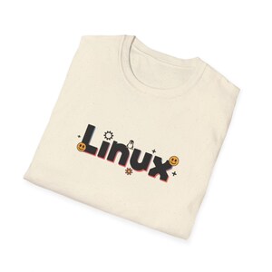 Puede incluir: Camiseta color crema con la palabra "Linux" en una fuente colorida y estilizada. El dise&ntilde;o incluye un engranaje, un ping&uuml;ino, una flor y caras sonrientes. La camiseta est&aacute; doblada.