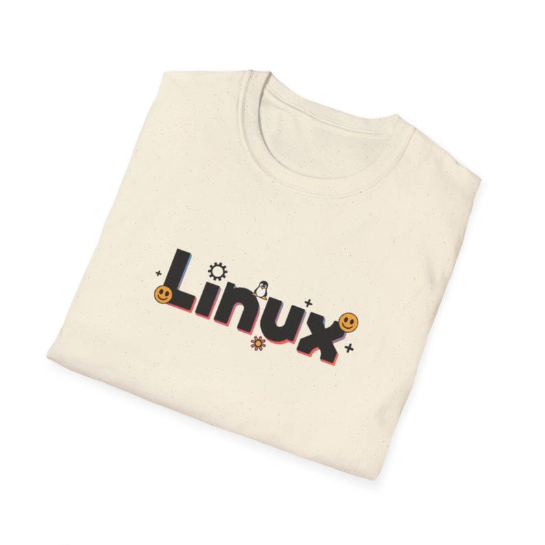 Puede incluir: Camiseta color crema con la palabra "Linux" en una fuente colorida y estilizada. El dise&ntilde;o incluye un engranaje, un ping&uuml;ino, una flor y caras sonrientes. La camiseta est&aacute; doblada.