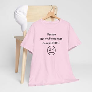 Puede incluir: Una camiseta rosa claro con el texto "Funny But not Funny HAHA Funny ERROR..." y un gr&aacute;fico de cara simple. Una gorra de b&eacute;isbol blanca cuelga de una silla de madera detr&aacute;s de la camiseta.