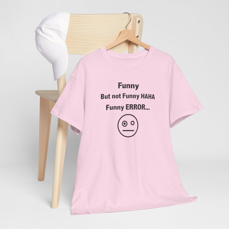 Puede incluir: Una camiseta rosa claro con el texto "Funny But not Funny HAHA Funny ERROR..." y un gr&aacute;fico de cara simple. Una gorra de b&eacute;isbol blanca cuelga de una silla de madera detr&aacute;s de la camiseta.