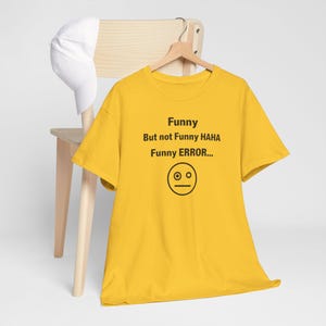 Puede incluir: Una camiseta amarilla con el texto "Funny But not Funny HAHA Funny ERROR..." y una cara con dos ojos y una l&iacute;nea recta para la boca. Una gorra de b&eacute;isbol blanca cuelga de una silla de madera.