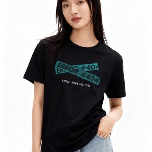 May include: Black crew neck t-shirt with a teal graphic that reads "ERROR #404 MIND NOT FOUND". The graphic is designed to look like crossed tape. The model is wearing blue jeans.