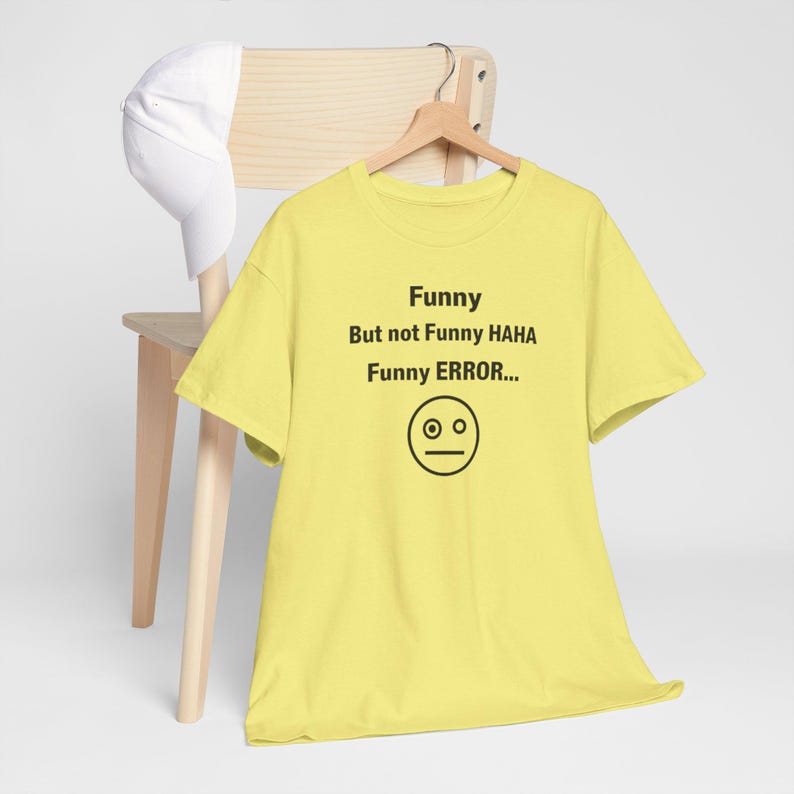 Puede incluir: Una camiseta amarilla con el texto "Funny But not Funny HAHA Funny ERROR..." y un gr&aacute;fico de cara simple. Una gorra de b&eacute;isbol blanca descansa sobre una silla de madera.
