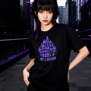 May include: Black t-shirt featuring a purple flame design with the text "LINUX FEELING NOT A DECISION". The short-sleeved shirt has a crew neck. The image is set outdoors with a city background.