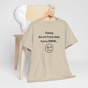 Puede incluir: Una camiseta beige con el texto "Funny But not Funny HAHA Funny ERROR..." y un gr&aacute;fico de cara simple. La camiseta est&aacute; colgada de una percha de madera, con una gorra de b&eacute;isbol blanca apoyada en una silla de madera.