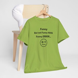 Puede incluir: Una camiseta verde lima con el texto "Funny But not Funny HAHA Funny ERROR..." y un gr&aacute;fico de cara simple. La camiseta est&aacute; colgada de una percha de madera, con una gorra de b&eacute;isbol blanca apoyada en una silla de madera en el fondo.