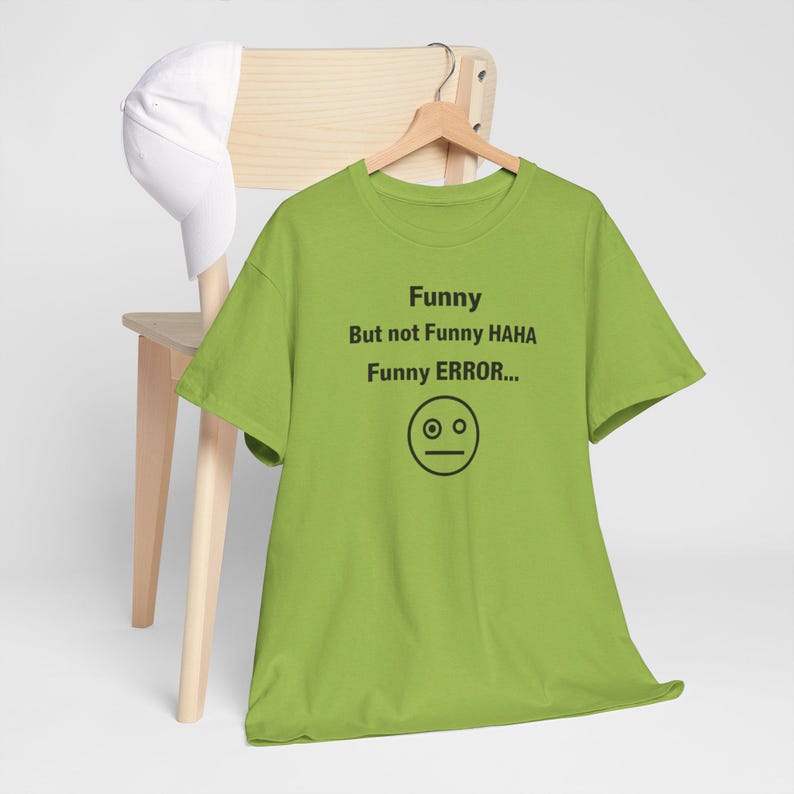 Puede incluir: Una camiseta verde lima con el texto "Funny But not Funny HAHA Funny ERROR..." y un gr&aacute;fico de cara simple. La camiseta est&aacute; colgada de una percha de madera, con una gorra de b&eacute;isbol blanca apoyada en una silla de madera en el fondo.