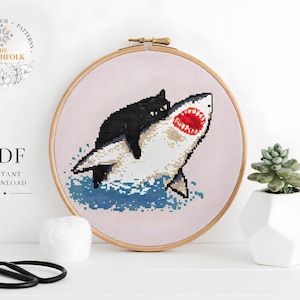 May include: Cross-stitch pattern of a black cat riding a shark, set in a wooden embroidery hoop. The shark has a wide-open mouth with red teeth. The text "PDF INSTANT DOWNLOAD" is visible.