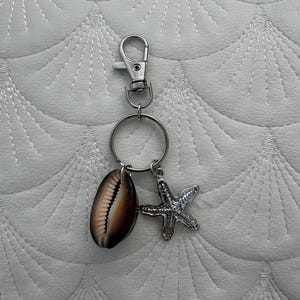 May include: A silver-toned metal keychain featuring a lobster clasp and a circular ring. Attached to the ring are two charms: a smooth, oval-shaped brown and cream cowrie shell and a textured, silver-toned metal starfish charm with five points.