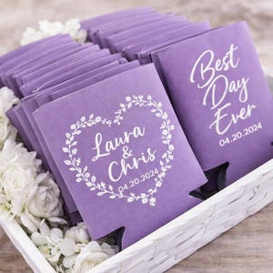 May include: Lavender-coloured can coolers, some with the text "Best Day Ever" and the date 04.20.2024, others with "Laura & Chris" inside a floral heart and the same date. The coolers are in a white woven basket.