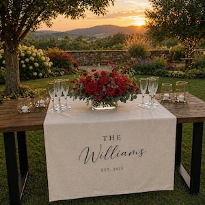 May include: A wooden table set for an outdoor event, featuring a cream-colored table runner with the text "The Williams" and "Est. 2025". A floral arrangement of red roses and greenery sits in a glass vase. Champagne flutes and candle holders are also on the table.