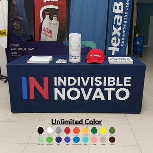 May include: A navy blue table cover with the text "INDIVISIBLE NOVATO" in white. A red baseball cap with the text "IND VISIBLE" is on the table. A stack of white paper and a stack of white cups are also on the table.