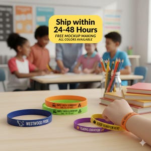 Könnte beinhalten: Eine Auswahl an bunten Silikonarmbändern mit Text. Die Bänder sind blau, grün, orange und lila. Der Text enthält "WESTWOOD PRIDE", "MAPLE CREEK ELEMENTARY", "READING CHAMPION" und "GOOD BEHAVIOR HERO". Der Hintergrund zeigt ein Klassenzimmer.