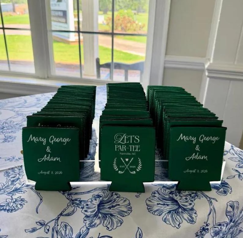 May include: A collection of dark green foam can coolers arranged in rows. Some feature the white printed text Mary George & Adam August 8 2026, while others display the text Let's Par-Tee Farmville, NC Est. 2026 with a golf club graphic.