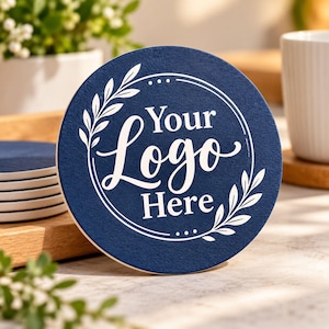 Puede incluir: Un posavasos circular de color azul marino con un diseño impreso en blanco de una corona de laurel que rodea el texto Your Logo Here. El posavasos tiene una superficie texturizada y forma parte de una pila sobre una bandeja de madera.