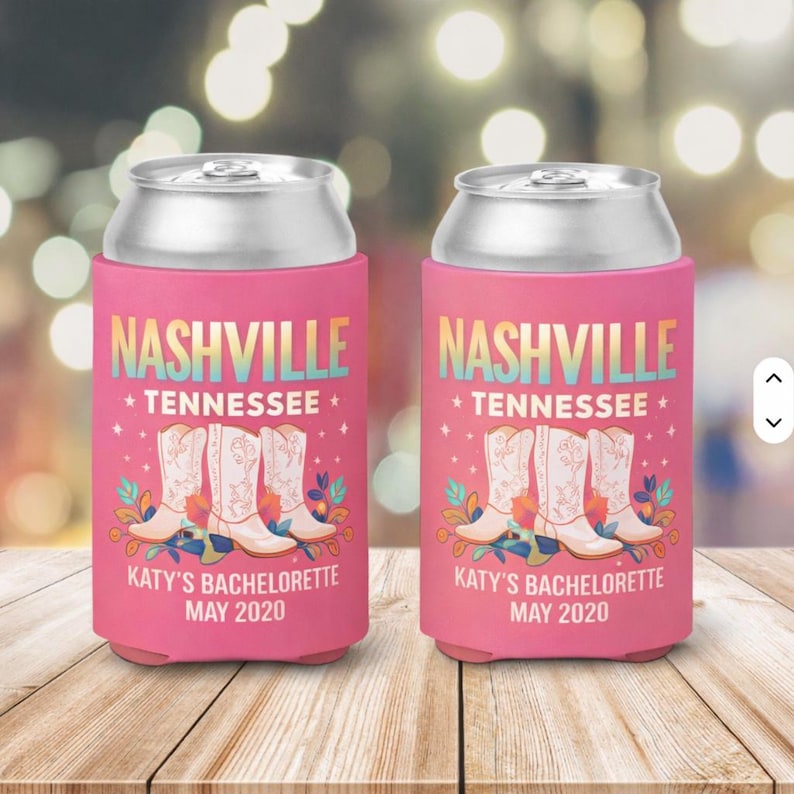 May include: Two pink foam can coolers featuring the text NASHVILLE TENNESSEE and KATY'S BACHELORETTE MAY 2020. The design includes three white cowboy boots surrounded by colourful floral accents on the front of each cylindrical beverage sleeve.