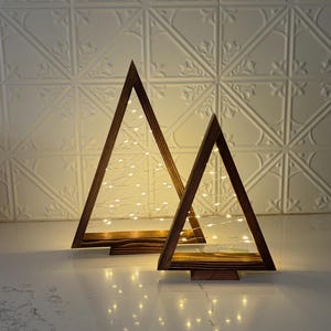 May include: Two wooden Christmas tree decorations with warm white fairy lights. The trees are triangular in shape and have a dark brown finish. The lights are strung inside the wooden frames, creating a cozy, festive glow. The trees are on a white surface.