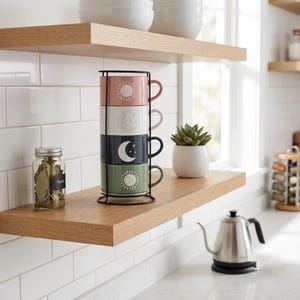 May include: A set of four stacked ceramic mugs in a black metal wire rack. The mugs feature celestial designs including suns and moons in shades of pink, white, navy blue, and sage green. Each mug has a side handle and a smooth, matte finish.
