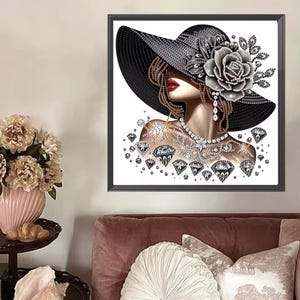 May include: A framed diamond painting art piece depicting a profile of a person wearing a wide-brimmed black hat adorned with a large silver rose. The figure features red lips, a silver necklace, and is surrounded by scattered diamond shapes.
