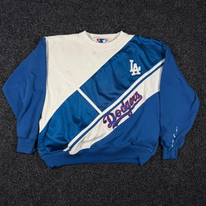 May include: A long-sleeve crewneck sweatshirt featuring a color-blocked design in royal blue and white. The front displays the Los Angeles Dodgers logo and the word Dodgers in script lettering. The garment has a ribbed collar, cuffs, and hem.