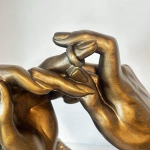 May include: Bronze-colored sculpture of two hands intertwined. The hands are detailed with visible knuckles and a textured surface, suggesting a metallic or stone material. The sculpture is set against a plain background.