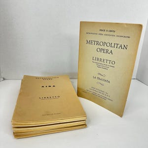 May include: Two vintage opera librettos, one titled "Aida" and the other "La Traviata." The "La Traviata" libretto is standing upright, and the "Aida" libretto is lying flat. Both are printed on aged, tan paper.