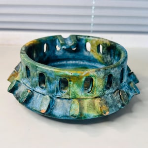 May include: A round ceramic ashtray featuring a mottled blue and green glaze with yellow highlights. The exterior wall has a series of vertical rectangular cutouts and a flared base with protruding rounded tabs around the entire circumference.