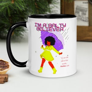Może przedstawiać: Biały kubek ceramiczny z czarnym rantem i uchwytem. Kubek przedstawia kolorową ilustrację osoby z parasolem i tekstem "I'm a Salty Believer". Zawiera również tekst "Ye are the salt of the world... Matthew 5:13".