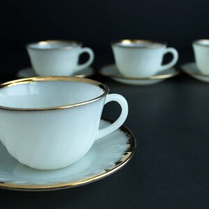 May include: Four white teacups with gold trim and matching saucers. The cups and saucers are arranged in a row on a black surface.