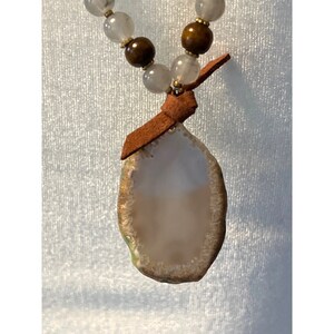 Peut inclure: Un collier en cordon de cuir marron avec un pendentif en agate de grande taille, de forme ovale. L'agate est translucide et présente un motif de couleur blanc et marron. Le collier est agrémenté de perles claires et brunes.