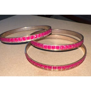 May include: Three circular bangles featuring a metallic gold-toned band inlaid with a continuous row of small, square-shaped, vibrant hot pink stones. The bracelets are arranged in a stacked formation, showcasing the reflective surface of the metal.