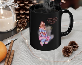 Puede incluir: Taza de café negra con una ilustración de una mujer en rosa y azul y el texto "Some things broke my heart but fixed my vision!"