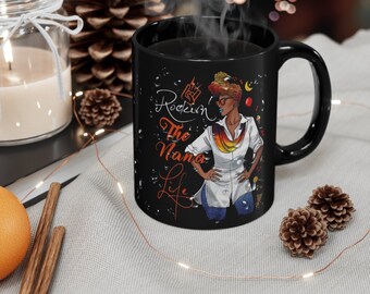 Puede incluir: Taza de café negra con una ilustración colorida de una mujer con afro, que lleva una bufanda colorida y pantalones vaqueros. El texto "Rockin' The Nana Life" está impreso en la taza.