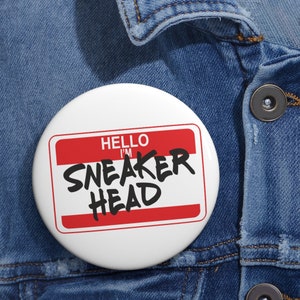 Puede incluir: Un botón blanco con una etiqueta de nombre roja que dice "Hello I'm Sneaker Head" en texto negro. El botón está sujeto a una chaqueta vaquera azul.