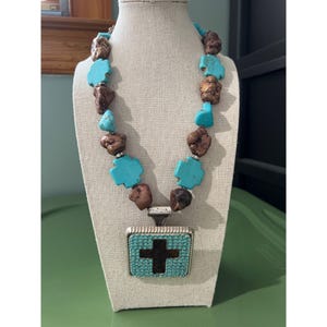 May include: A necklace featuring alternating turquoise-colored cross-shaped beads and irregular brown textured beads. The centerpiece is a square pendant with a silver-toned frame, a border of small light blue stones, and a dark brown cross inlay.