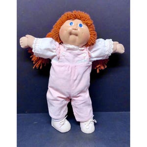 May include: A doll with orange yarn hair and blue eyes wearing a white shirt with lace-trimmed sleeves under light pink corduroy overalls. The doll has a round face, a small nose, and is wearing white shoes with laces tied in a bow at the front.
