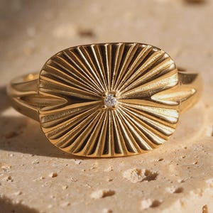 May include: A gold ring featuring an oval-shaped face with a sunburst pattern of radiating lines. A single, small, clear round gemstone is set in the center of the sunburst design. The band of the ring is smooth and tapers toward the back.