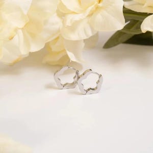 May include: A pair of silver-toned hoop earrings featuring a distinct five-petaled flower shape. The earrings have a polished metallic finish and a hinged closure mechanism, designed as small circular hoops with scalloped edges forming a floral outline.